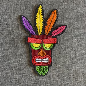 May include: Embroidered patch of a tribal mask with a red and brown face, green eyes, and a white-toothed mouth. The mask has colourful feathers in yellow, orange, pink, and purple. The patch has a black border.