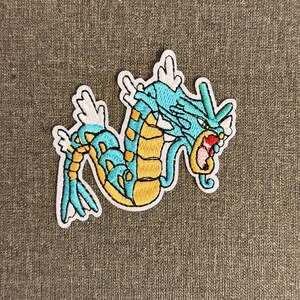 May include: Embroidered patch featuring a stylized aquatic dragon in shades of blue, yellow, and white. The dragon has a long, serpentine body with fins and a detailed head. The patch is outlined in white.