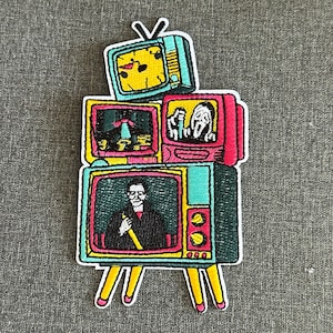 May include: Embroidered patch depicting a stack of vintage televisions. Each screen shows a different image, including a clock, a scene with a figure, and a person holding a yellow object. The patch has yellow legs and a white border.