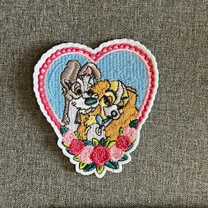 Op de afbeelding: Geborduurde patch met twee honden in een hartvormig ontwerp. De patch heeft een lichtblauwe achtergrond met een roze rand en een witte omtrek. De honden zijn omgeven door roze en groene bloemaccenten.