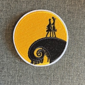 May include: A circular patch with a silhouette of two figures on a hill, set against a yellow moon. The hill and the moon's border are white, with a black spiral design. Suitable for clothing or accessories.