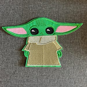 Peut inclure: Écusson brodé représentant un personnage vert avec de grandes oreilles, des yeux noirs et une robe beige. Le patch présente un design détaillé avec des accents roses sur les oreilles, pour vêtements ou accessoires.
