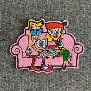 May include: Embroidered patch featuring a cartoon family on a pink couch. The characters have distinct features: blonde hair, red hair, and blue goggles. The patch has a pink border and is likely for clothing or accessories.