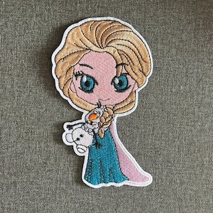 Op de afbeelding: Geborduurde patch met een cartoonafbeelding van een personage met blond haar, blauwe ogen en een turquoise jurk, die een sneeuwpop vasthoudt. De patch heeft een witte rand en is geplaatst tegen een grijze achtergrond.