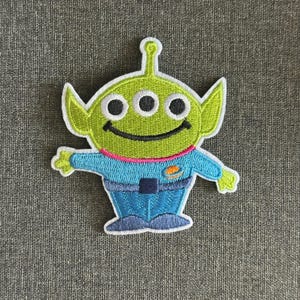 May include: Embroidered patch depicting a green alien with three eyes, large ears, and a smiling mouth. The alien is wearing a blue uniform with a pink collar and an orange emblem. The patch has a white border, suitable for clothing or accessories.