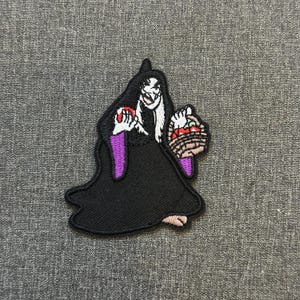Può includere: Toppa ricamata raffigurante una strega dei cartoni animati con mantello e cappuccio neri, che tiene in mano una mela rossa e un cestino di frutta. La strega ha i capelli bianchi e un sorriso sinistro, con accenti viola sulle maniche. Sfondo grigio.