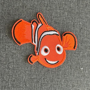 May include: Embroidered orange clownfish patch with white and black accents. The patch features a smiling face and is likely inspired by a popular animated film. This patch is suitable for clothing or accessories.