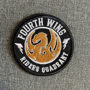Puede incluir: Un parche circular negro con texto blanco que dice "FOURTH WING" y "RIDERS QUADRANT". El centro presenta un diseño de fénix dorado. Dos rayos blancos flanquean el diseño. El parche es probablemente para ropa o accesorios.