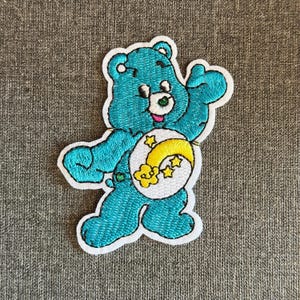 Pode incluir: Patch bordado com um urso azul-turquesa com contorno branco. O urso tem um rosto sorridente e um emblema na barriga com uma lua crescente amarela e estrelas. Fundo de tecido cinza.