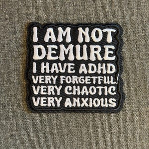Puede incluir: Un parche negro con texto bordado en blanco que dice "I AM NOT DEMURE I HAVE ADHD VERY FORGETFUL VERY CHAOTIC VERY ANXIOUS". El parche tiene un borde negro y está sobre una tela gris.
