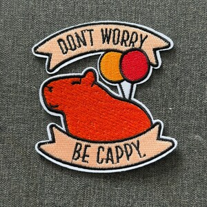 Peut inclure: Un écusson brodé représentant un capybara orange avec deux ballons, un jaune et un rouge. L'écusson comprend deux bannières pêche clair avec le texte noir DONT WORRY BE CAPPY. Le motif présente une bordure blanche et des coutures noires.
