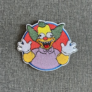 May include: Embroidered patch featuring Krusty the Clown from The Simpsons. The patch shows Krusty with a yellow face, green hair, and a purple shirt, set against a red circle with a blue border. His arms are outstretched.