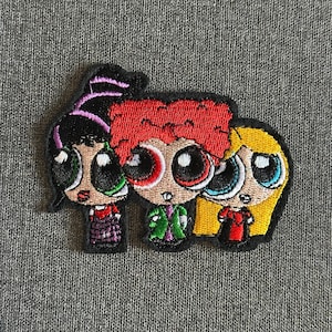 Pode incluir: Patch bordado com três personagens de desenhos animados com olhos grandes e penteados distintos. Os personagens têm cabelo preto, vermelho e amarelo e usam roupas de várias cores. O patch está em um fundo de tecido cinza.