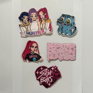 May include: A selection of embroidered patches with cartoon designs and text. The patches depict a group of three women with "HUNTR/X", a blue cat, a woman with red hair and "2IMEWOJ", a pink rectangle with "KPOP DEMON HUNTERS", and a heart with "Sait Boys".