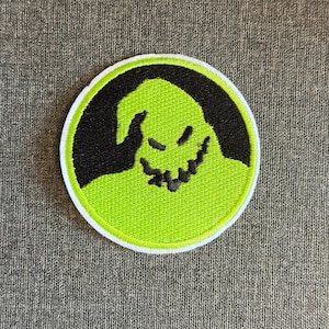 May include: A circular patch with a green character design, black outline, and a grinning face. The character is set against a black background within a white-bordered circle. Suitable for clothing or accessories.