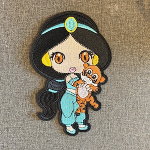 Puede incluir: Parche bordado con un personaje de dibujos animados con cabello negro, que lleva un atuendo turquesa y sostiene un pequeño tigre naranja. El parche tiene un borde negro y está sobre un fondo gris.