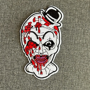 Può includere: Toppa ricamata raffigurante una faccia da clown spaventosa con dettagli di sangue rosso e un cappello a bombetta nero. Il clown ha una faccia bianca, un'espressione sinistra e un ampio sorriso sanguinante. La toppa ha un bordo bianco.
