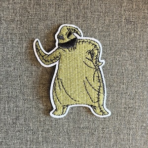 Könnte beinhalten: Gestickter Aufnäher mit einer Figur in Grün und Schwarz, mit einem genähten Design und weißer Umrandung. Die Figur hat einen gebogenen Arm erhoben. Der Aufnäher ist für das Aufnähen auf Kleidung oder Accessoires gedacht.