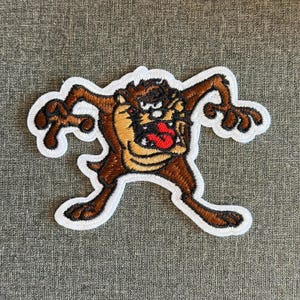 May include: Embroidered patch featuring the Tasmanian Devil cartoon character. The patch has a white border and depicts the character in shades of brown, tan, and red, with an open mouth and a dynamic pose.