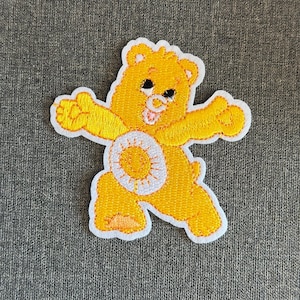 May include: Embroidered yellow bear patch with a white border. The bear has a smiling face, outstretched arms, and a white circle with a yellow center on its belly. The patch is designed to be ironed or sewn onto fabric.