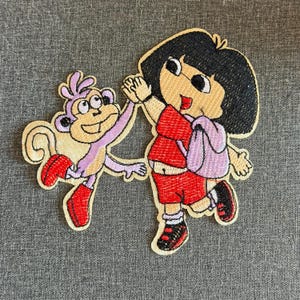 Op de afbeelding: Geborduurde patch met Dora de Explorer en Boots de aap die een high five geven. Dora draagt een rode outfit en een paarse rugzak. Boots is paars met rode laarzen. De patch heeft een gele rand.