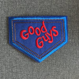 Può includere: Una toppa blu con la scritta "Good Guys" in rosso. La toppa ha la forma di un piatto di casa da baseball con un bordo cucito marrone e un contorno blu. Su uno sfondo di tessuto grigio strutturato.