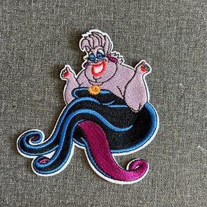 Könnte beinhalten: Gestickter Aufnäher mit Ursula aus Arielle, die Meerjungfrau. Der Aufnäher zeigt Ursula mit lila Haut, einem schwarzen Kleid und blauen und lila Tentakeln. Der Aufnäher hat einen weißen Rand.