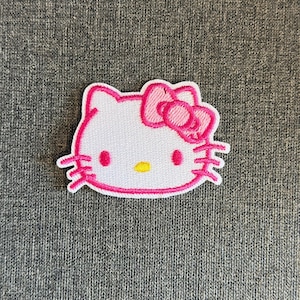 Könnte beinhalten: Gestickter Hello Kitty Aufnäher. Der Aufnäher hat ein weißes Gesicht mit rosa Umrandung, Schleife und Details. Die Augen sind rosa und die Nase gelb. Ideal zum Anpassen von Kleidung oder Accessoires. Ungefähr 5 cm hoch.
