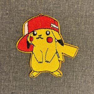 May include: Embroidered patch of a yellow Pikachu character wearing a red baseball cap. The patch has black, white, and red accents, with a detailed stitched design. The character has red cheeks and a grumpy expression.