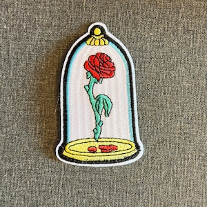 Può includere: Toppa ricamata con una rosa rossa all'interno di una cupola di vetro. La rosa ha foglie e stelo verdi, su una base gialla con contorno nero. La toppa è bianca con bordo blu e parte superiore gialla.