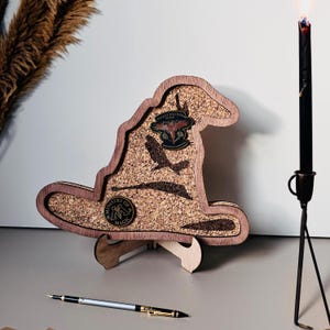 May include: A wooden Harry Potter Sorting Hat-shaped display piece with a corkboard interior. The hat features a dark brown frame, a corkboard center, and decorative metal accents. The base is made of light-coloured wood. A black candle is lit in the background.