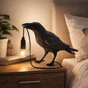 May include: A dark grey crow-shaped lamp with a light bulb held in its beak. The lamp sits on a wooden bedside table. The lamp's black cord forms a loop on the table. The bulb emits a warm glow.