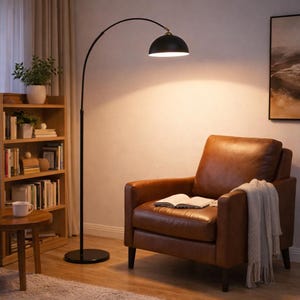 Può includere: Una lampada da terra ad arco nera con un accento dorato, che illumina una poltrona in pelle marrone. Un libro e una coperta sono sulla sedia. Una libreria in legno e un tavolino sono anch'essi visibili.