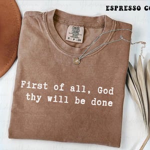 Könnte beinhalten: Espressofarbenes T-Shirt mit dem weißen Text "First of all, God thy will be done". Das Shirt hat einen Rundhalsausschnitt und ein kleines Etikett. Eine silberne Halskette mit Anhänger und getrockneten Blättern ist ebenfalls zu sehen.
