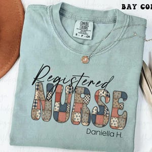 Puede incluir: Camiseta azul claro con la frase "Registered Nurse" en un diseño de patchwork. El nombre "Daniella H." también está impreso debajo. Se ve un collar plateado con un colgante.