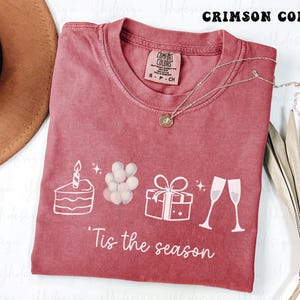 Puede incluir: Camiseta de color carmesí con ilustraciones gráficas blancas de una tarta, globos, un regalo y copas de champán. El texto "'Tis the season" está impreso debajo. Un collar dorado está sobre la camiseta.