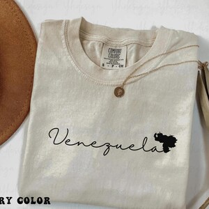 Puede incluir: Camiseta de color marfil con la palabra "Venezuela" en cursiva y una silueta negra del país. Un collar dorado con un colgante está drapeado sobre la camiseta. Un sombrero marrón es visible en el fondo.