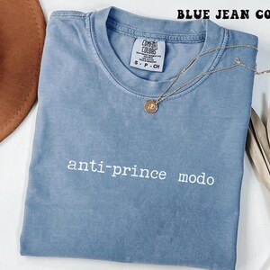 Puede incluir: Una camiseta Comfort Colors azul vaquero con el texto "anti-prince modo" en blanco. Un collar dorado con un colgante descansa sobre la camiseta. La camiseta está doblada sobre una superficie blanca.