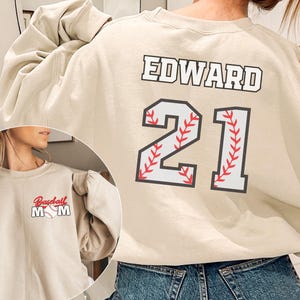 Puede incluir: Una sudadera beige con el nombre "EDWARD" y el número "21" impreso en la espalda. El número está estilizado como una pelota de béisbol con costuras rojas. La parte delantera de la sudadera tiene una pelota de béisbol con las palabras "Baseball Mom" impresas.