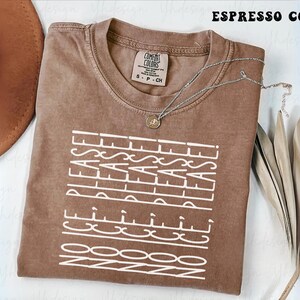 Puede incluir: Camiseta color espresso con el texto "NO ICE PLEASE!" en blanco. La camiseta tiene cuello redondo y una pequeña etiqueta en el cuello. También se ve un collar plateado con un colgante y una planta seca.