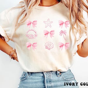 Puede incluir: Camiseta color marfil con la palabra "IVORY COLOR" en la parte inferior. Presenta diseños rosas de lazos, una estrella de mar, una concha, una palmera y un atardecer. Los elementos son de color rosa claro.
