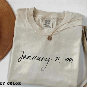 Puede incluir: Camiseta Comfort Colors color marfil con el texto "January 21, 1991" en escritura negra. Un collar dorado con un colgante descansa sobre la camiseta. Un sombrero marrón es visible en la esquina superior izquierda.