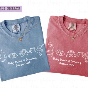 Puede incluir: Dos camisetas, una azul y otra roja, con diseños gráficos y texto en blanco. El texto de las camisetas dice "Baby Olivia is Growing October 2026". Cada camiseta tiene un collar dorado. Las camisetas están etiquetadas como "Couple Shirts".