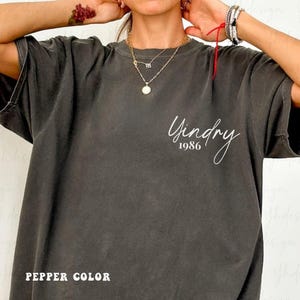 Puede incluir: Camiseta de gran tamaño color pimienta con mangas cortas. La camiseta presenta el texto "Yindry 1986" en cursiva en la parte delantera. La camiseta es de color gris oscuro.