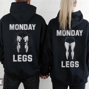 Op de afbeelding: Zwarte hoodies met de tekst "MONDAY LEGS" en een afbeelding van benen in wit en grijs. De afbeelding toont de achterkant van de hoodies, met het woord "BLACK" onderaan.