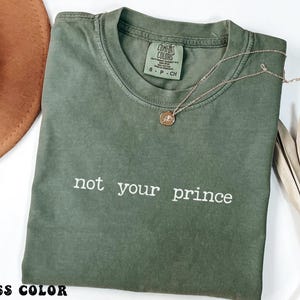 Puede incluir: Una camiseta de color musgo con la frase "not your prince" impresa en blanco. Un collar dorado con un colgante descansa sobre la camiseta. Un sombrero marrón y hojas secas son visibles en el fondo.