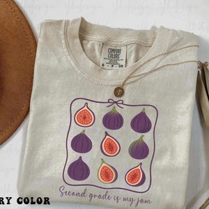Op de afbeelding: Ivoor Comfort Colors T-shirt met een vijgenafbeelding. De afbeelding toont negen vijgen, sommige heel en sommige gesneden, in een paarse lijst. De tekst "Second grade is my jam" staat eronder. Een gouden ketting ligt op het T-shirt.