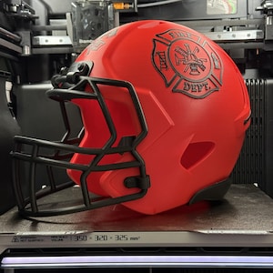 Peut inclure: Casque de football américain rouge avec un masque noir et l'emblème des pompiers. Le casque est dans une imprimante 3D. Le volume de construction est de 350 x 320 x 325 mm³.