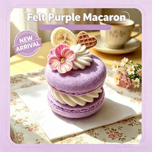 Peut inclure: Un macaron en feutrine violette surmonté d'un tourbillon en feutrine blanche, d'une fleur en tissu rose, d'une tranche de banane en feutrine et d'une petite gaufre en forme de cœur marron. Le texte "Felt Purple Macaron" et "NEW ARRIVAL" est visible.