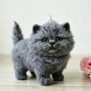 Puede incluir: Una figura de gatito gris de lana fieltrada con un pelaje texturizado y esponjoso, grandes ojos oscuros y largos bigotes blancos. El pequeño gato está de pie en posición frontal con la cola levantada, junto a flores rosas.